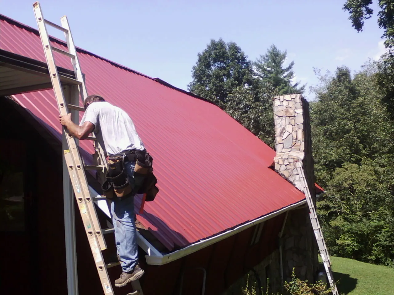 Roofer on a ladder beside a red metal roof and stone chimney — Soffit & Fascia Repair by Roofing Brothers in Mc Dermott